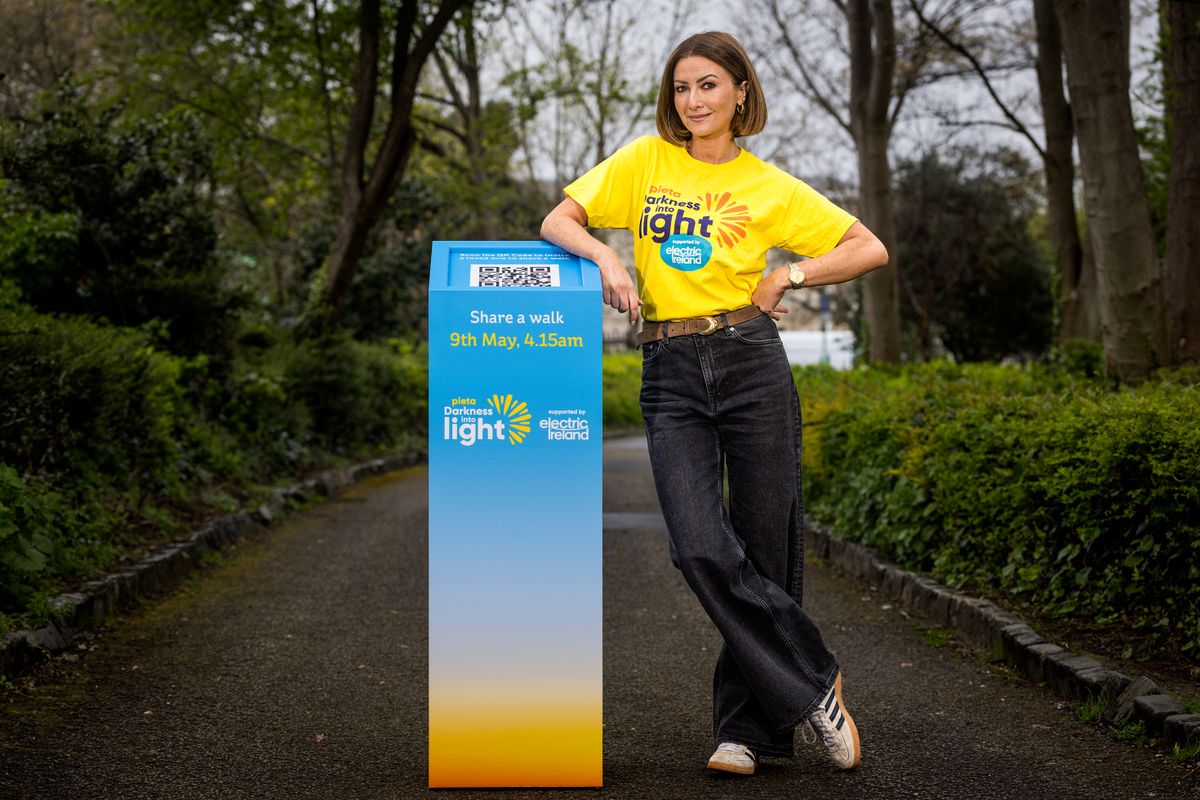 Woman in a yellow "Darkness Into Light" shirt stands by a sign for a walk event on 9th May at 4:15 AM, set in a park.